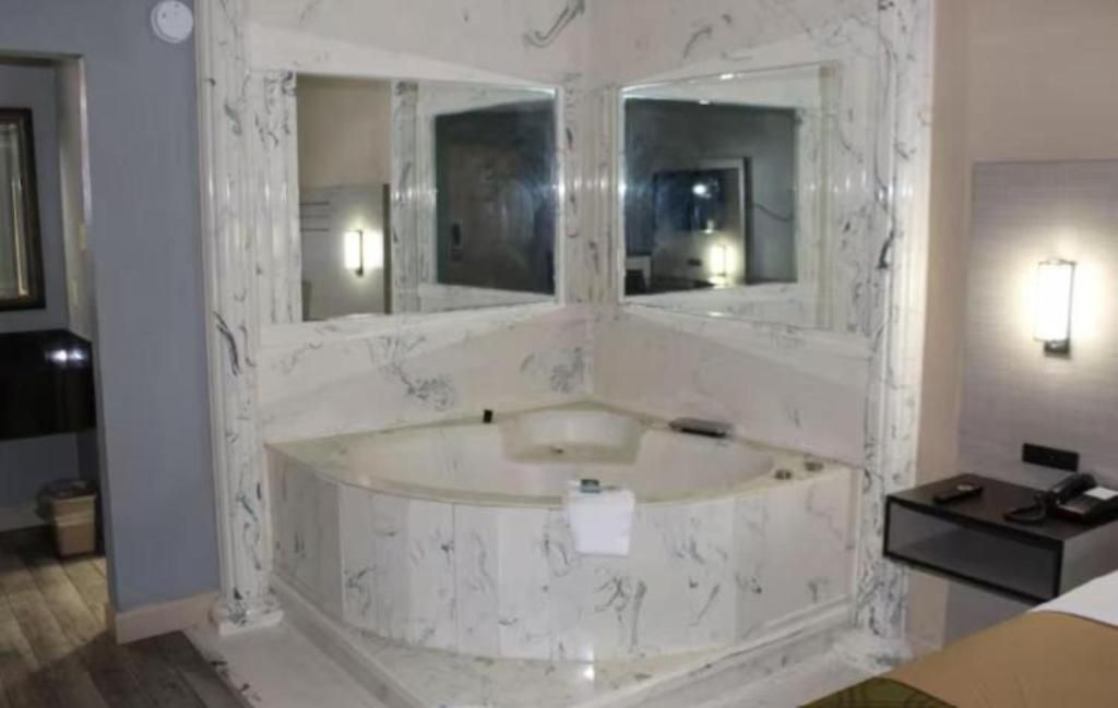 A bathroom with a marble bathtub, two wall-mounted mirrors, and a small bedside table with a lamp.