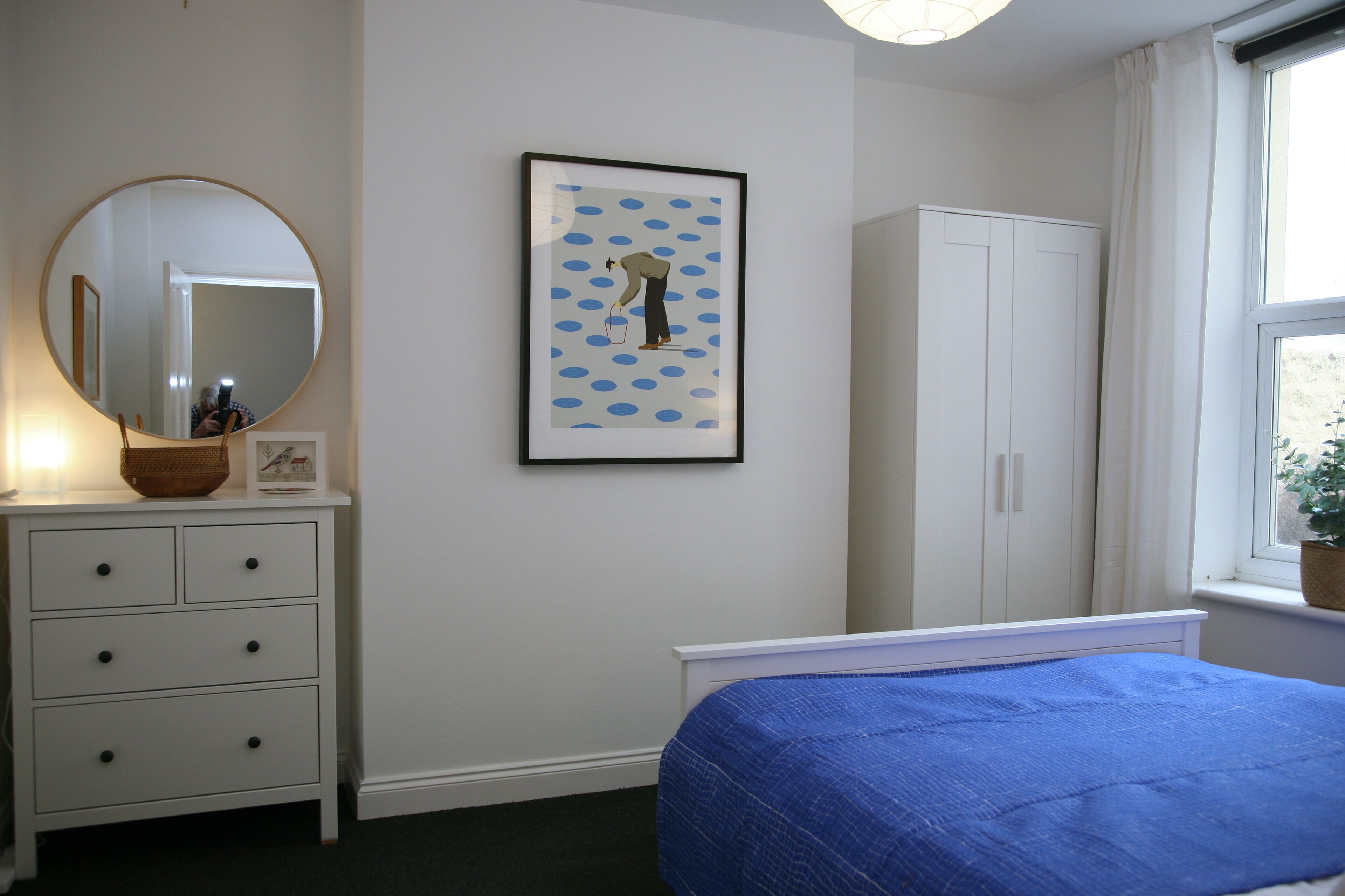 Une chambre à coucher avec un lit, une commode, un miroir, un tableau encadré et une fenêtre avec une plante.