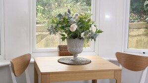 A corner room with a wooden table and two chairs, a vase with flowers, and a view of greenery through a window. - Charming one bed  flat near Brighton Station and short walk to North Laine (Brighton)