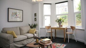 A cozy living room with a sofa, chairs, a small table, and a bookshelf. There are large windows with a view of the outside. - Charming one bed  flat near Brighton Station and short walk to North Laine (Brighton)