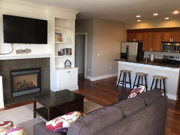 A modern living room with a fireplace, a flat-screen TV, a sofa, and a kitchen area with a refrigerator, microwave, and stools.