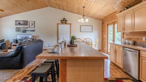 Un interior de cabaña espacioso con techo de madera, un comedor con mesa larga, una cocina con armarios de madera y una zona de estar acogedora con sofá y sillones.