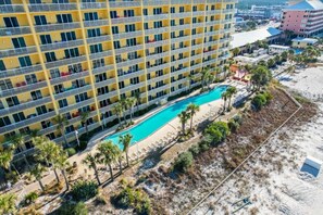Hotel features a distinctive facade, balconies with seating, and a pool area surrounded by a sandy beach and palm trees.