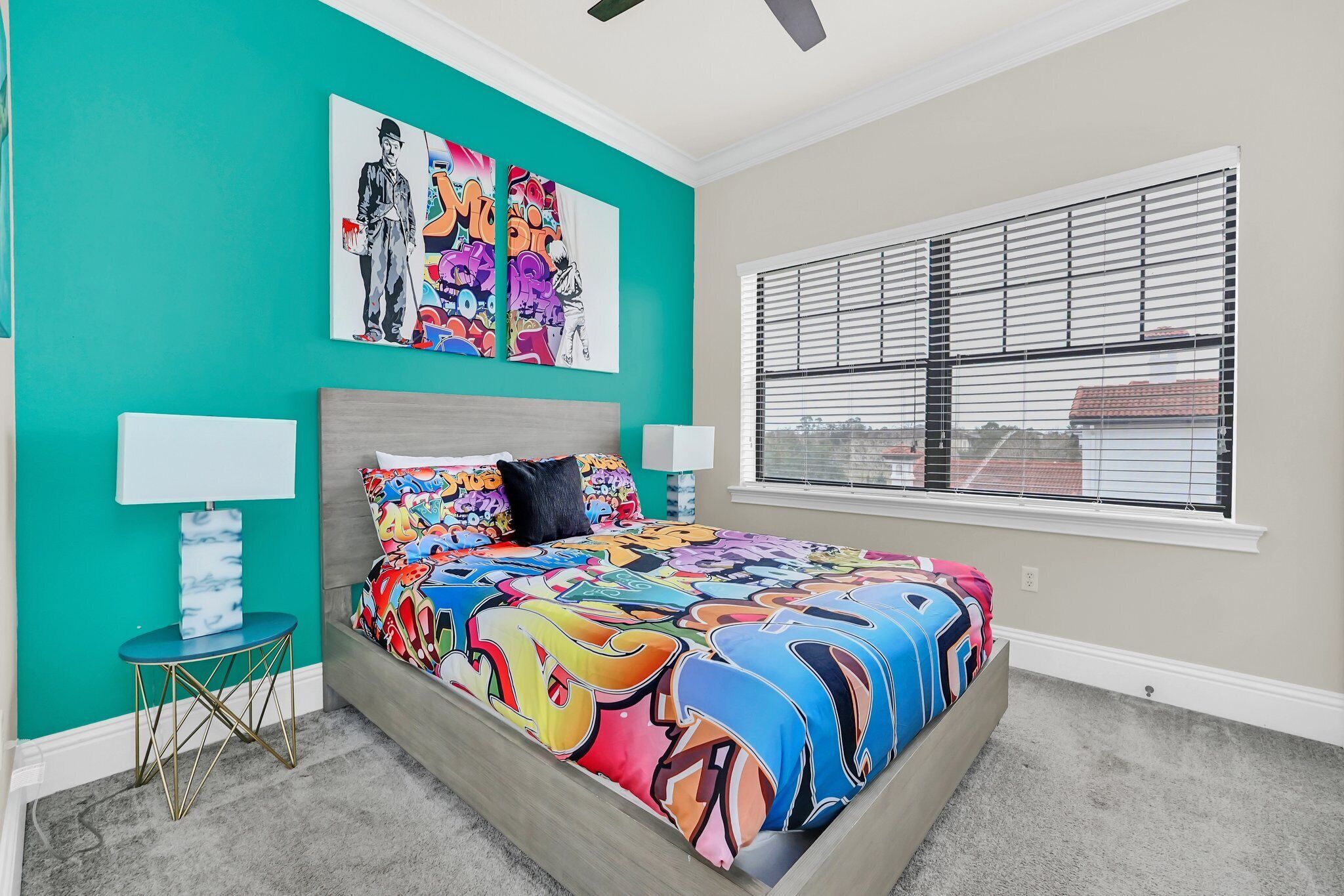A bedroom with a bed, a window with blinds, a small table with a lamp, and a wall with two pieces of artwork.