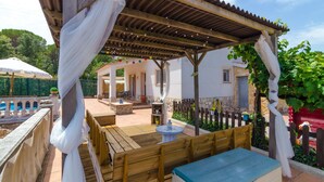 A covered outdoor seating area with wooden benches and a table, a stone wall, and a building with a tiled roof in the background. - Club Villamar - Nice house, complete and very sunny, located in a quiet urbanization,with a  priv... (Vidreres)