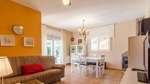 A living room with a sofa, a glass coffee table, a dining area with a white table and chairs, a chandelier, and a kitchen area with a stove. - Club Villamar - Nice house, complete and very sunny, located in a quiet urbanization,with a  priv... (Vidreres)