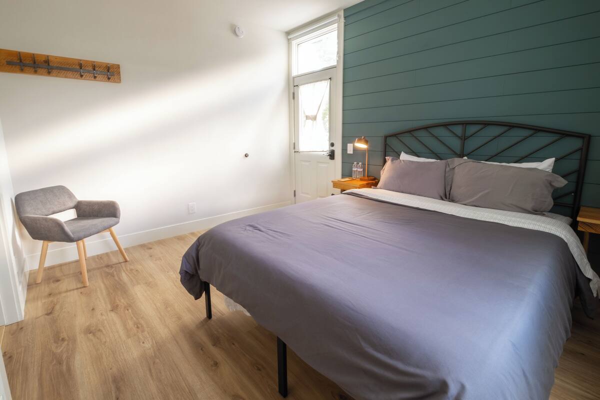 A bedroom with a bed, a chair, a nightstand with a lamp, and a window.