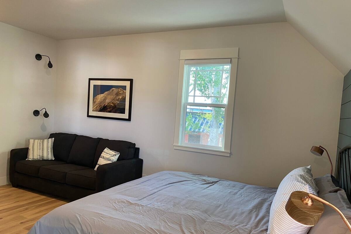 A bedroom with a bed, a dark-colored sofa, a window with a view of greenery, and a framed picture on the wall.