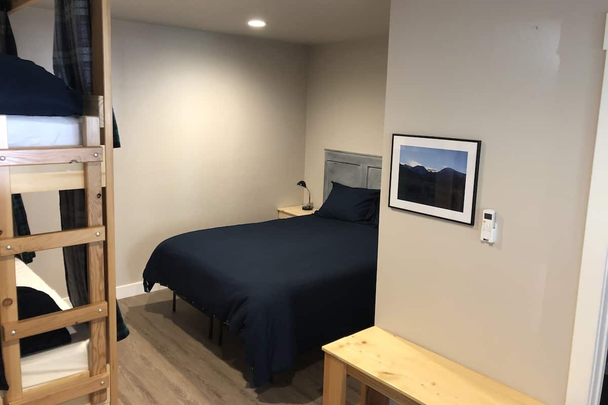 A bedroom with a bunk bed, a single bed, a bedside table with a lamp, a framed picture on the wall, and a small wooden bench.