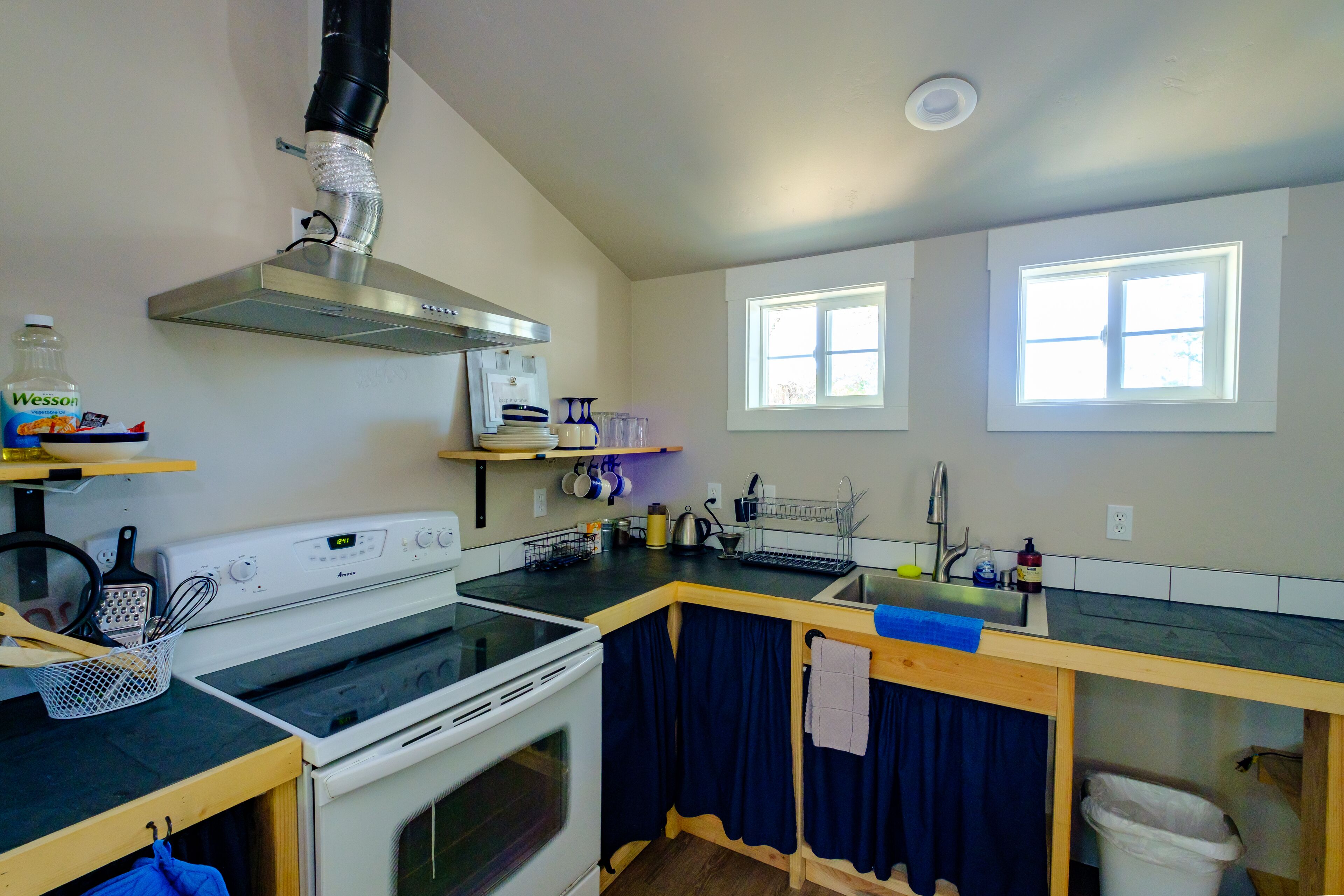 Cozinha compacta com fogão, forno e exaustor de aço inoxidável. Há duas janelas que oferecem luz natural, e uma pequena lixeira é visível.