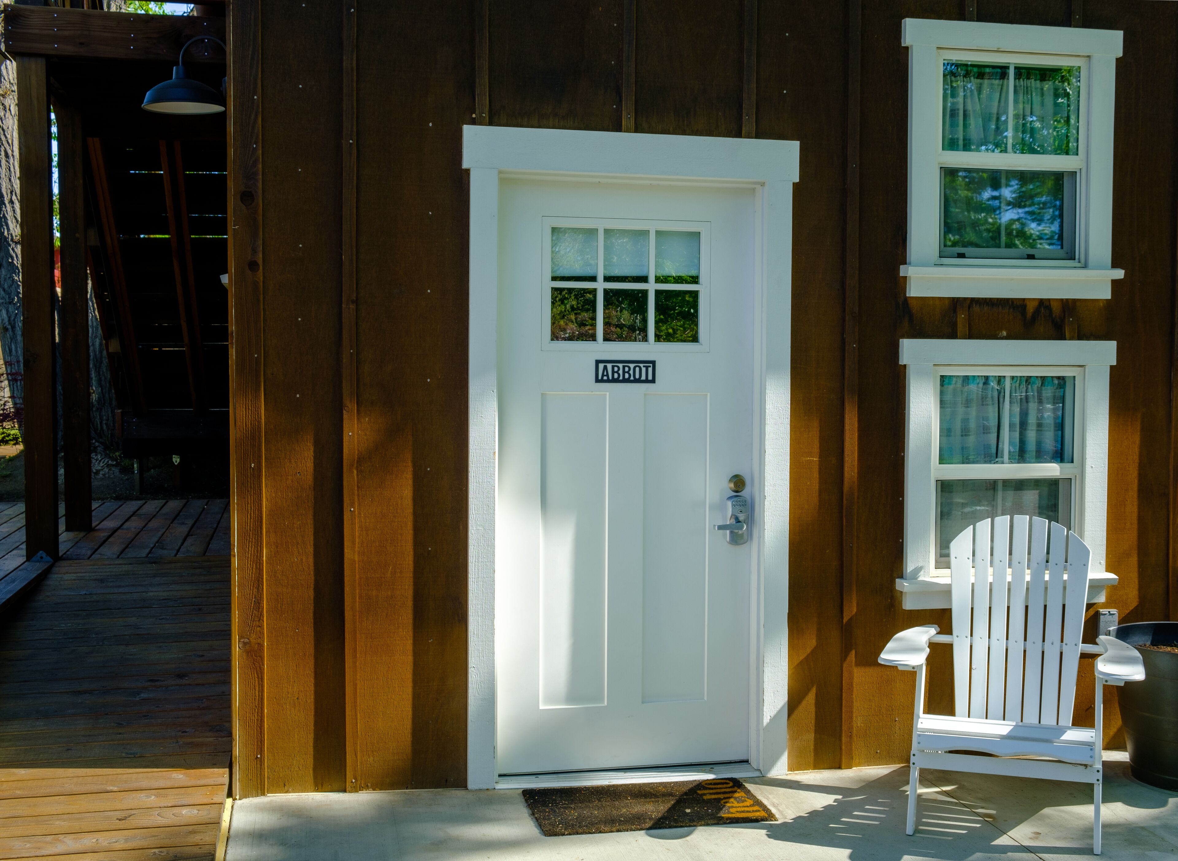 A wooden cabin with a white door labeled "ABBOT", a white chair outside, and a small window with curtains.