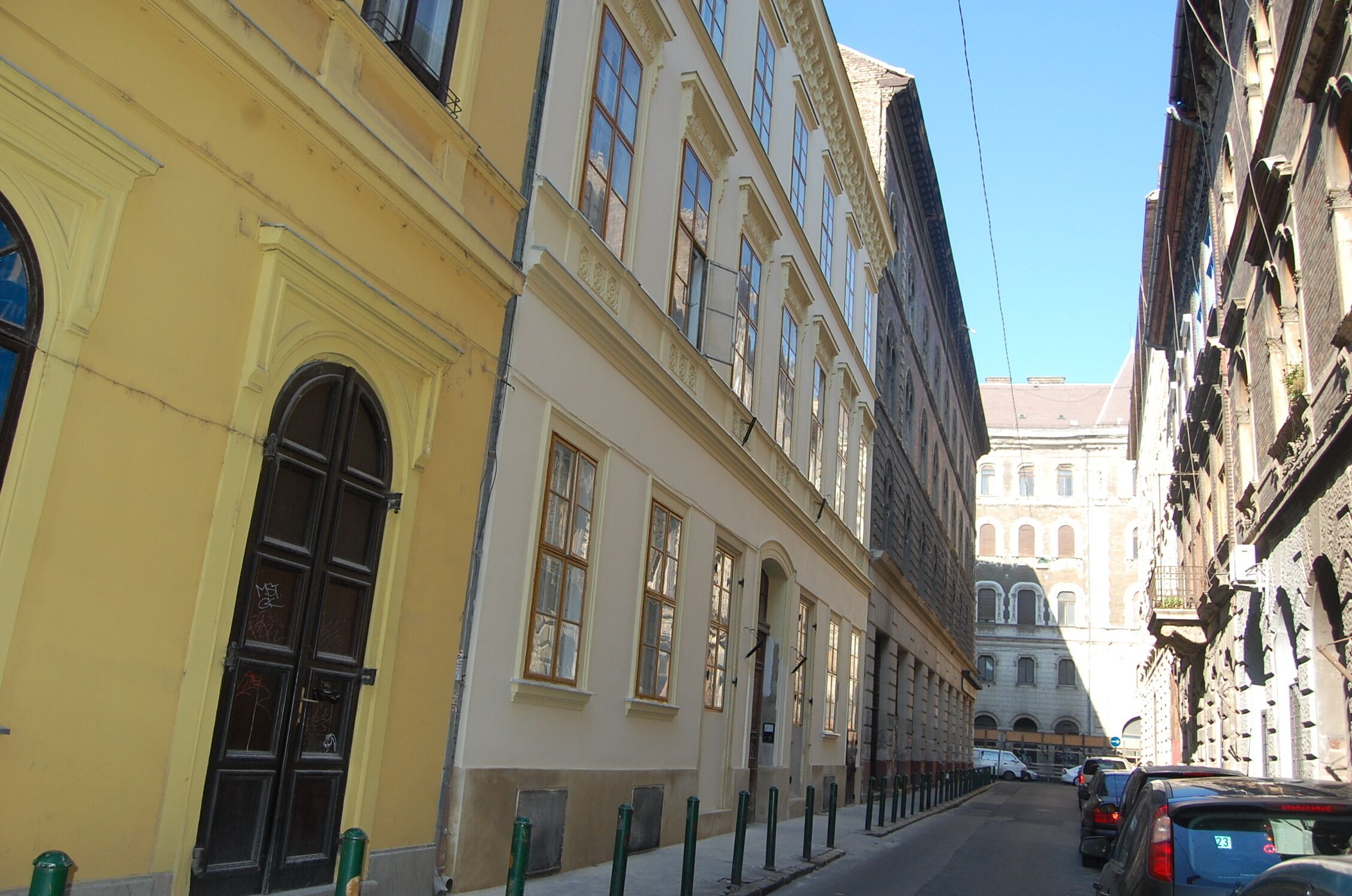 A narrow street with historic buildings, a prominent yellow building with a large arched entrance, and a clear blue sky.