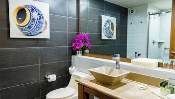 Un baño moderno con baldosas oscuras, un inodoro blanco, un lavabo color crema y un espejo que refleja un jarrón azul y blanco.