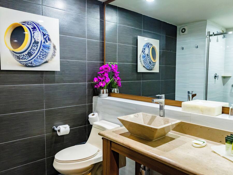 Un baño moderno con baldosas oscuras, un inodoro blanco, un lavabo color crema y un espejo que refleja un jarrón azul y blanco.