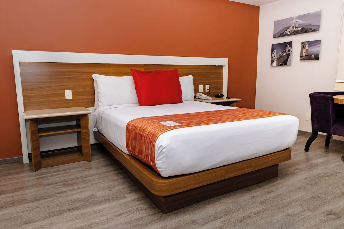 A hotel room with a wooden headboard, a bed with white and orange bedding, a nightstand with a phone, and a desk with a chair.