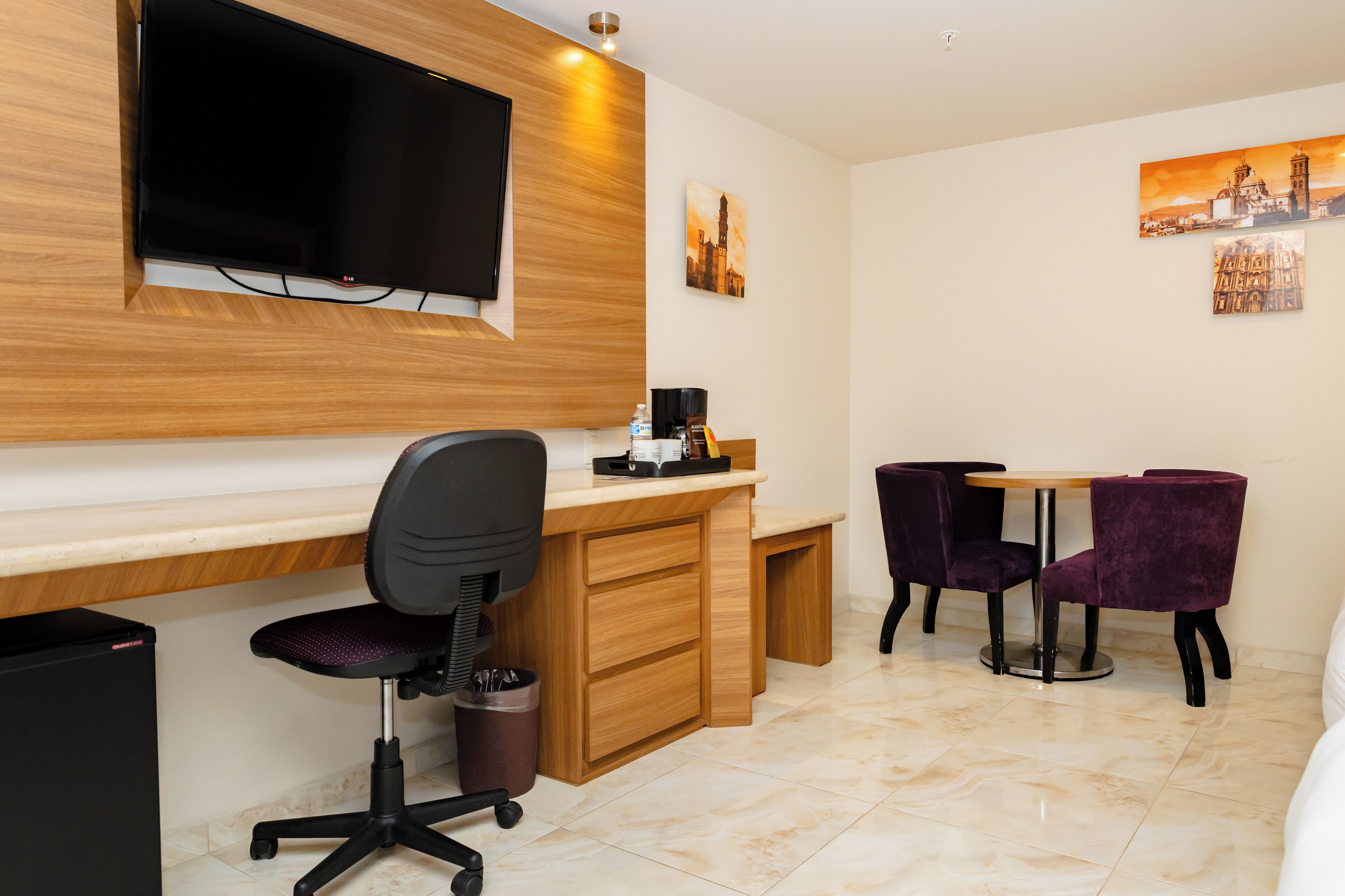 A modern hotel room with a wooden desk, a black office chair, a small table with purple chairs, a flat-screen TV mounted on the wall, and artwork depicting cityscapes.