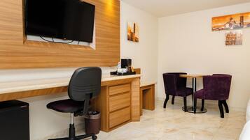 A modern hotel room with a wooden desk, a black office chair, a small table with purple chairs, a flat-screen TV mounted on the wall, and artwork depicting cityscapes.