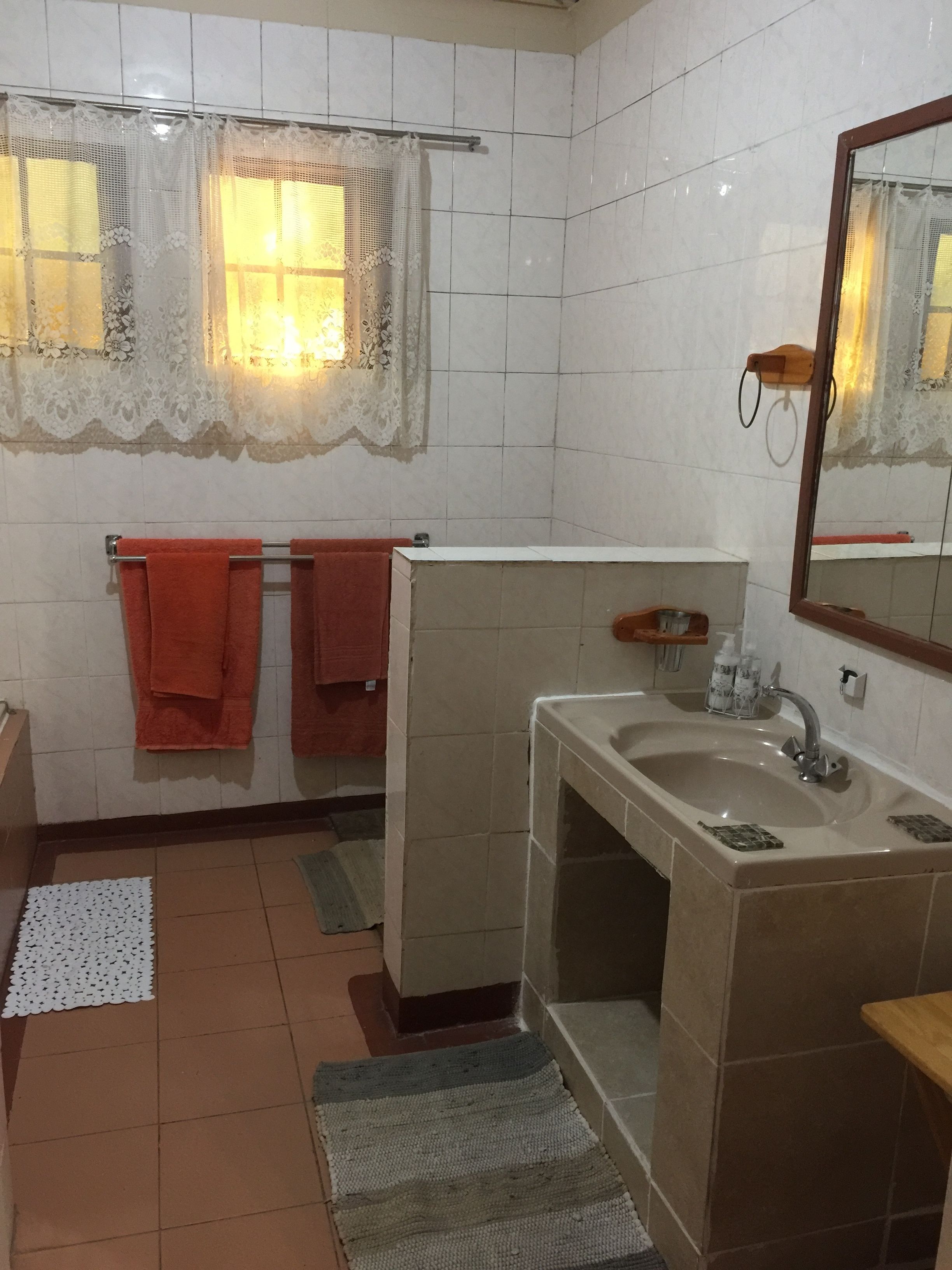 Un baño con paredes de azulejos blancos, un lavamanos con espejo, dos toallas rojas, una ventana con cortinas de encaje y un estante de madera pequeño con artículos de aseo.