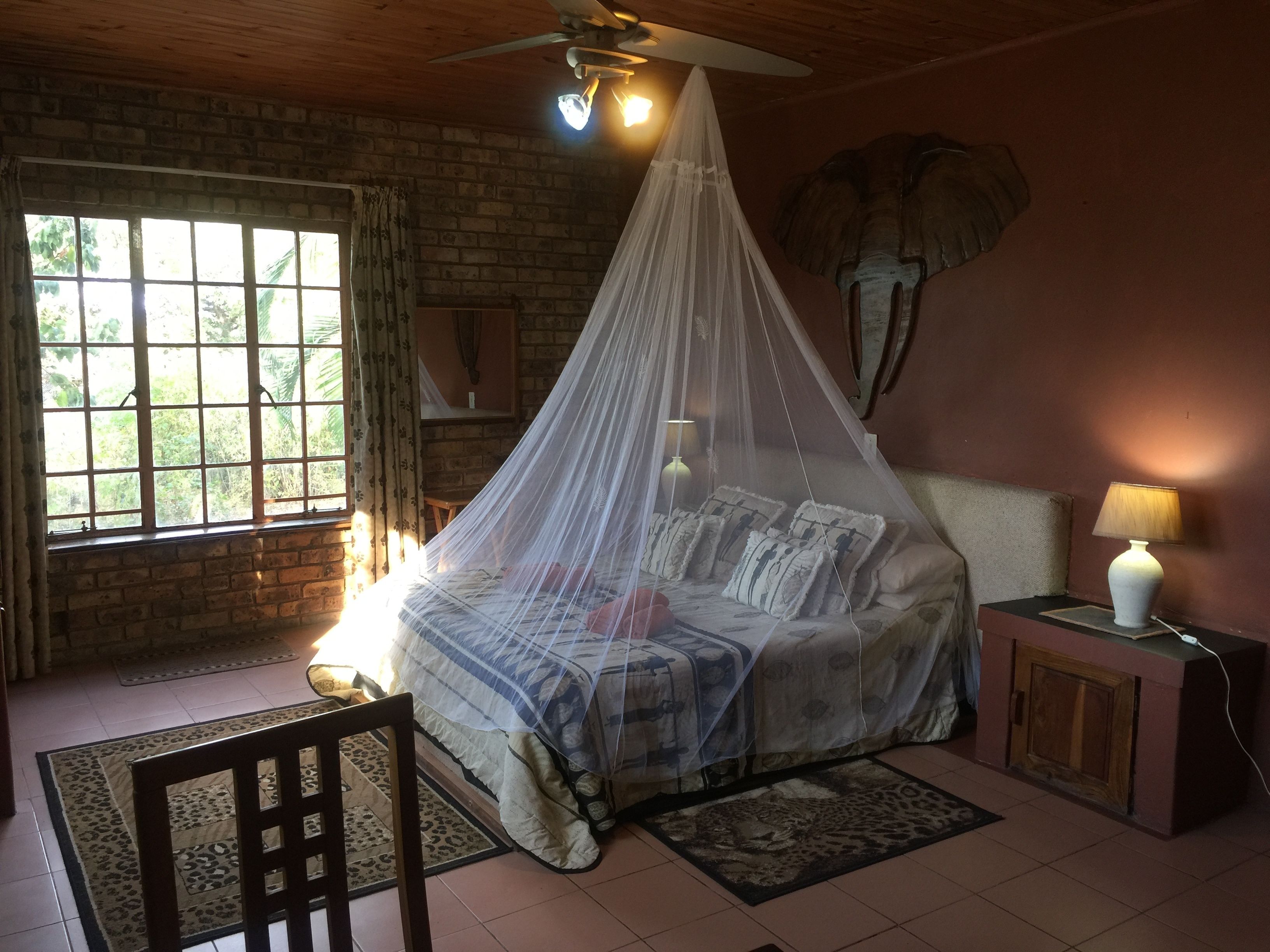 A bedroom with a bed under a mosquito net, a nightstand with a lamp, a chair, a window with curtains, and a brick wall.
