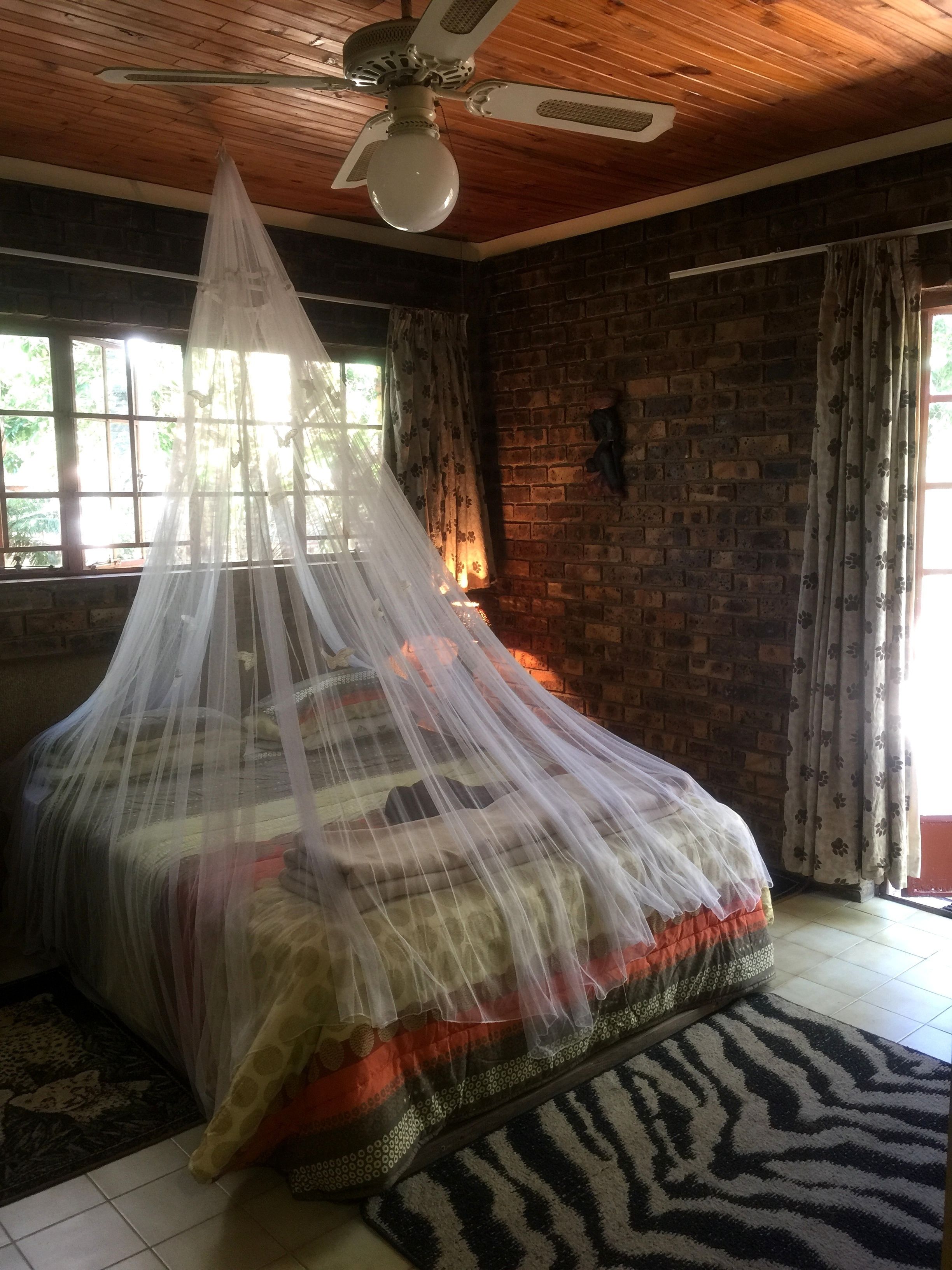 A room with a bed covered by a mosquito net, a wooden ceiling fan, brick walls, and a window with curtains.