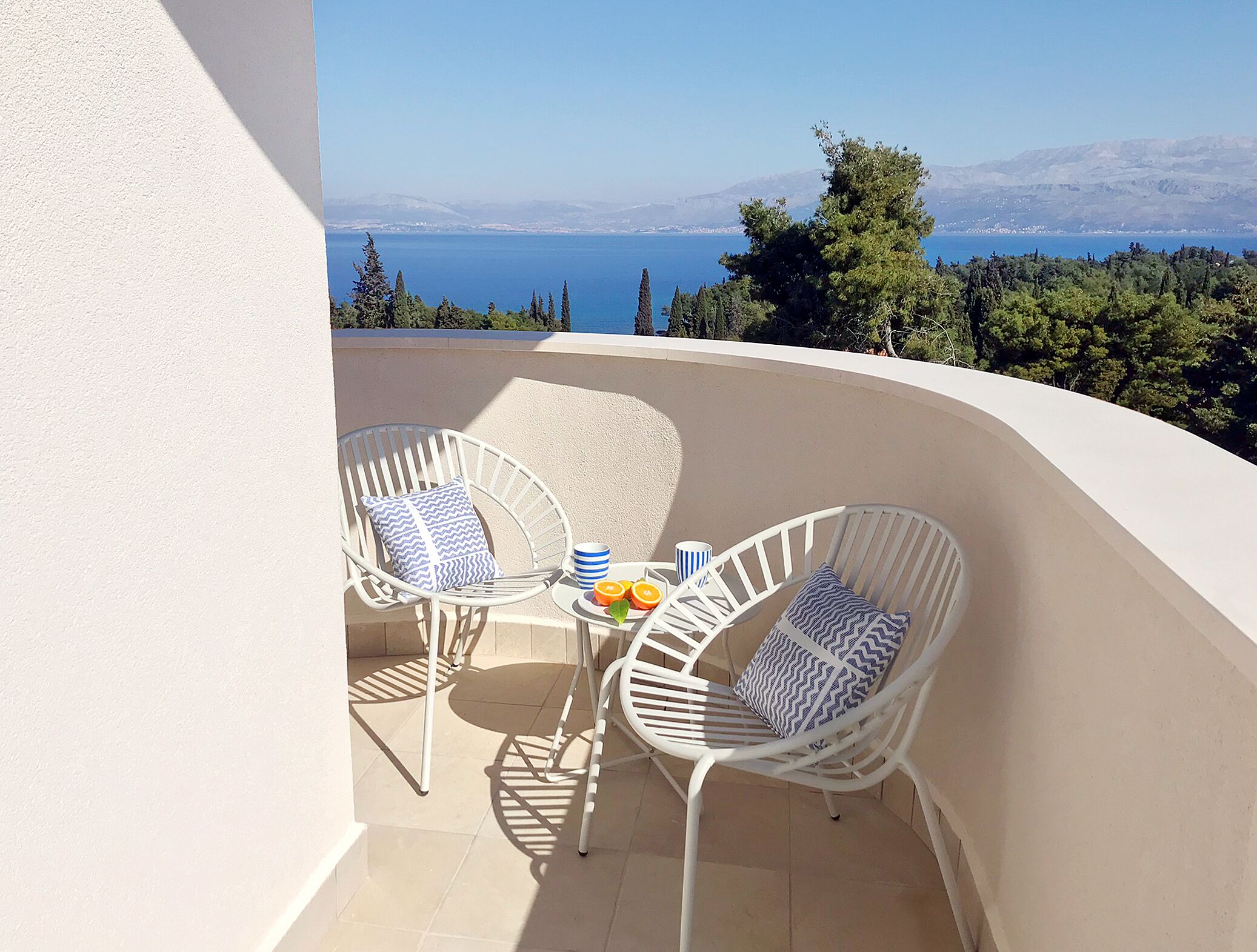A balcony with two white wicker chairs and a small round table, overlooking a scenic view of the sea and distant mountains.
