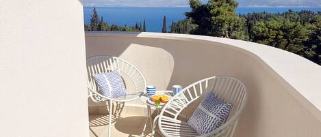 A balcony with two white wicker chairs and a small round table, overlooking a scenic view of the sea and distant mountains.