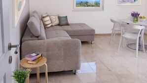Un salon moderne avec un canapé d’angle gris, une petite table ronde sur laquelle sont posés des livres, un vase avec des fleurs et un coin repas comprenant une table et des chaises blanches.