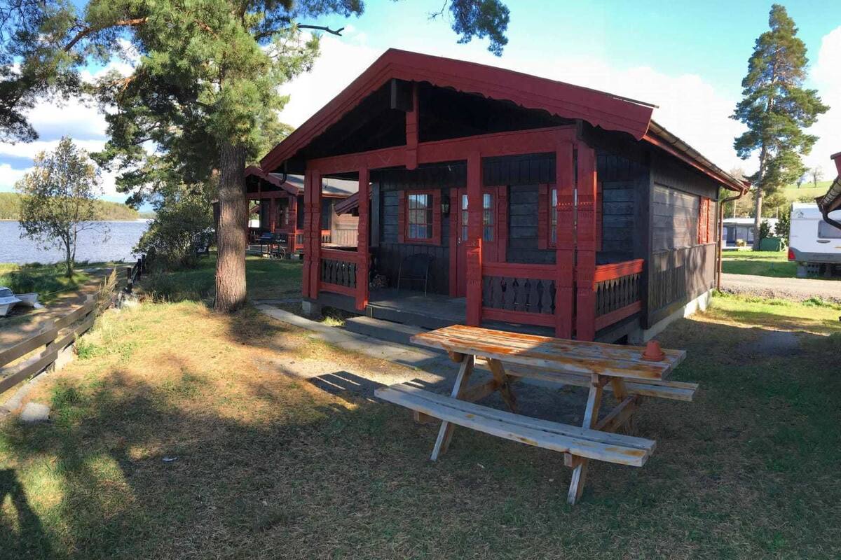 A rustic cabin with a red roof and wooden walls, a picnic table in front, and a lake in the background.