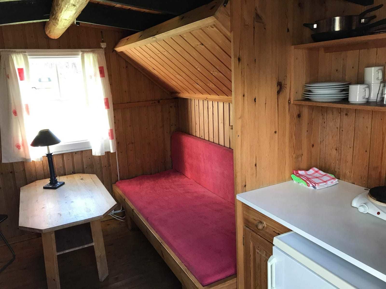 Una habitación tipo cabaña compacta con interior de madera, una cama pequeña con colchón rojo, una mesa blanca con lámpara y una kitchenette con estufa y repisas.