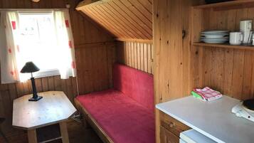 Una habitación tipo cabaña compacta con interior de madera, una cama pequeña con colchón rojo, una mesa blanca con lámpara y una kitchenette con estufa y repisas.