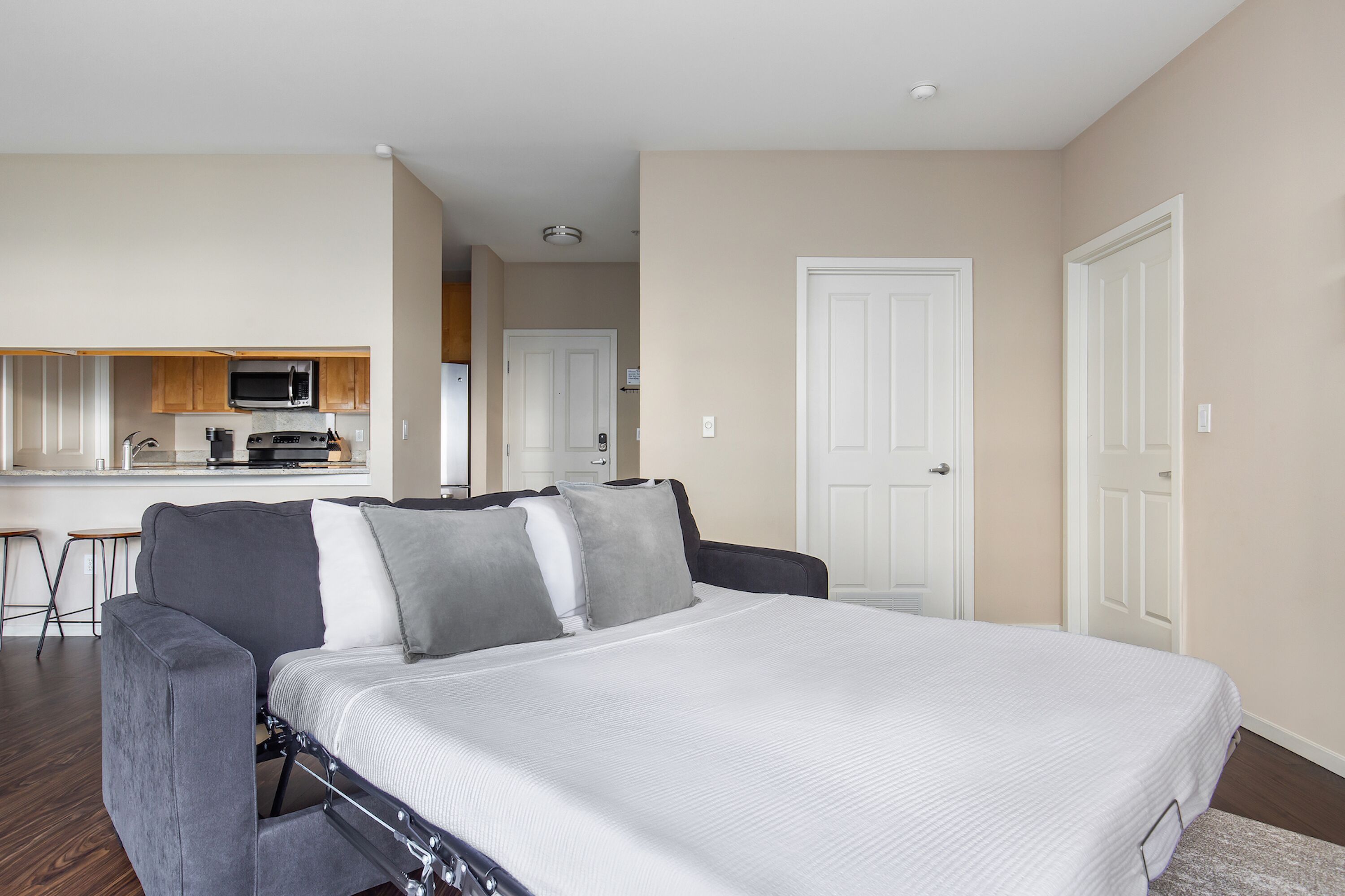 A bedroom with a bed, a gray couch, a kitchenette with a microwave, and a white door.