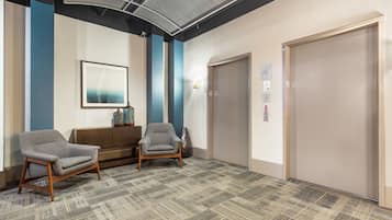 A waiting area with two grey armchairs, a wooden bench, a framed picture on the wall, and an elevator.