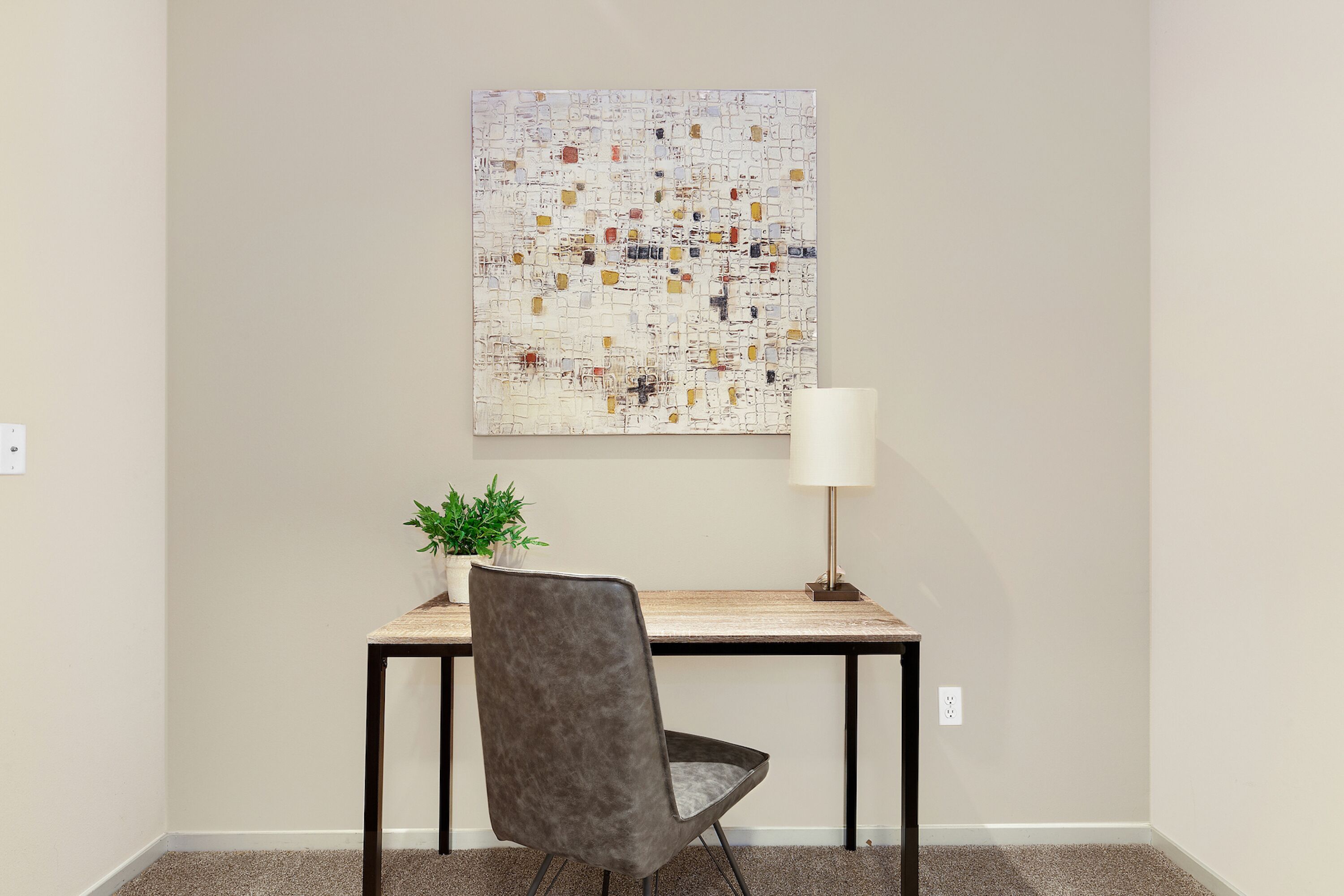 A room with a desk, chair, lamp, and a wall-mounted abstract painting.