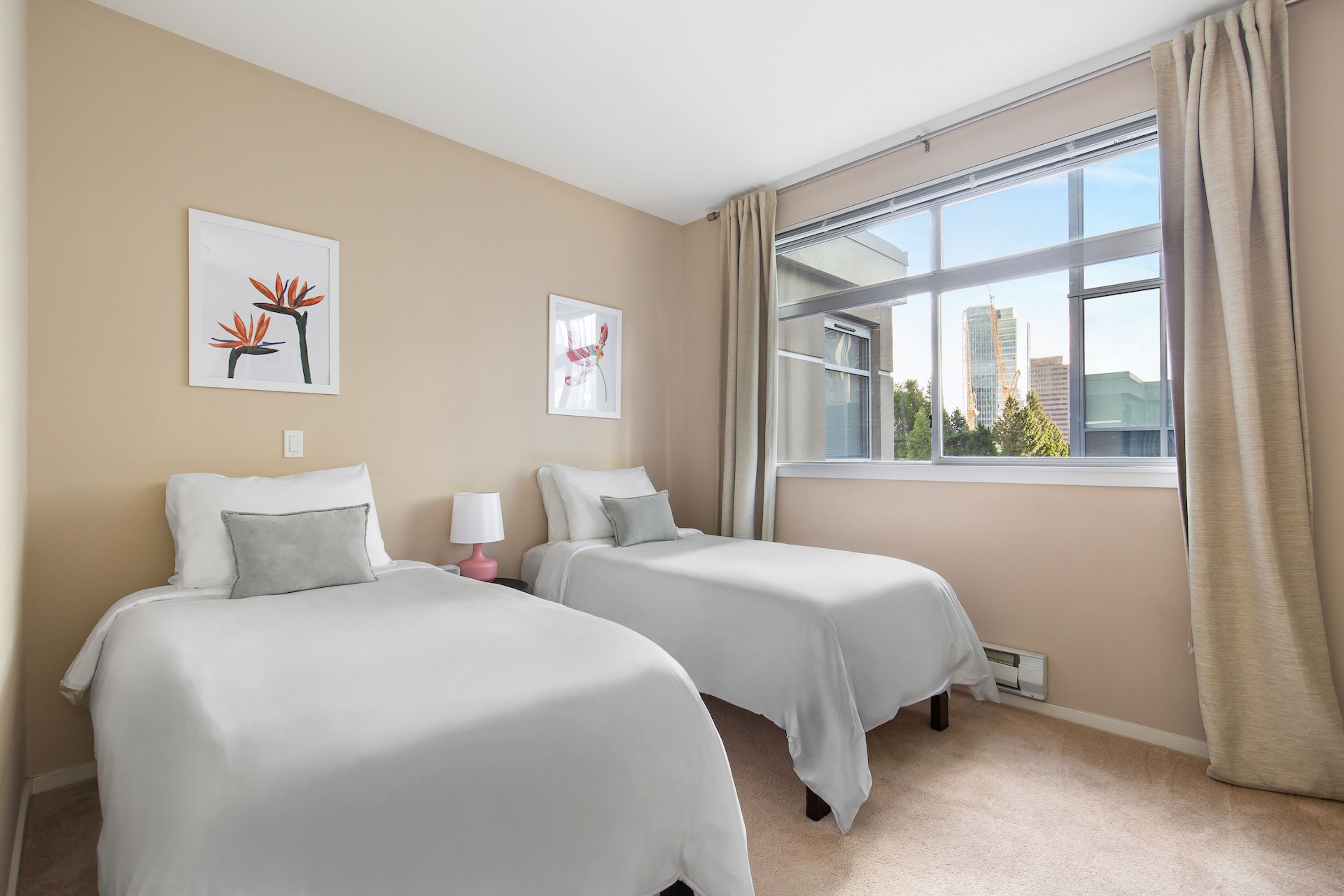 A hotel room with two beds, beige walls, and a large window with a view of buildings.