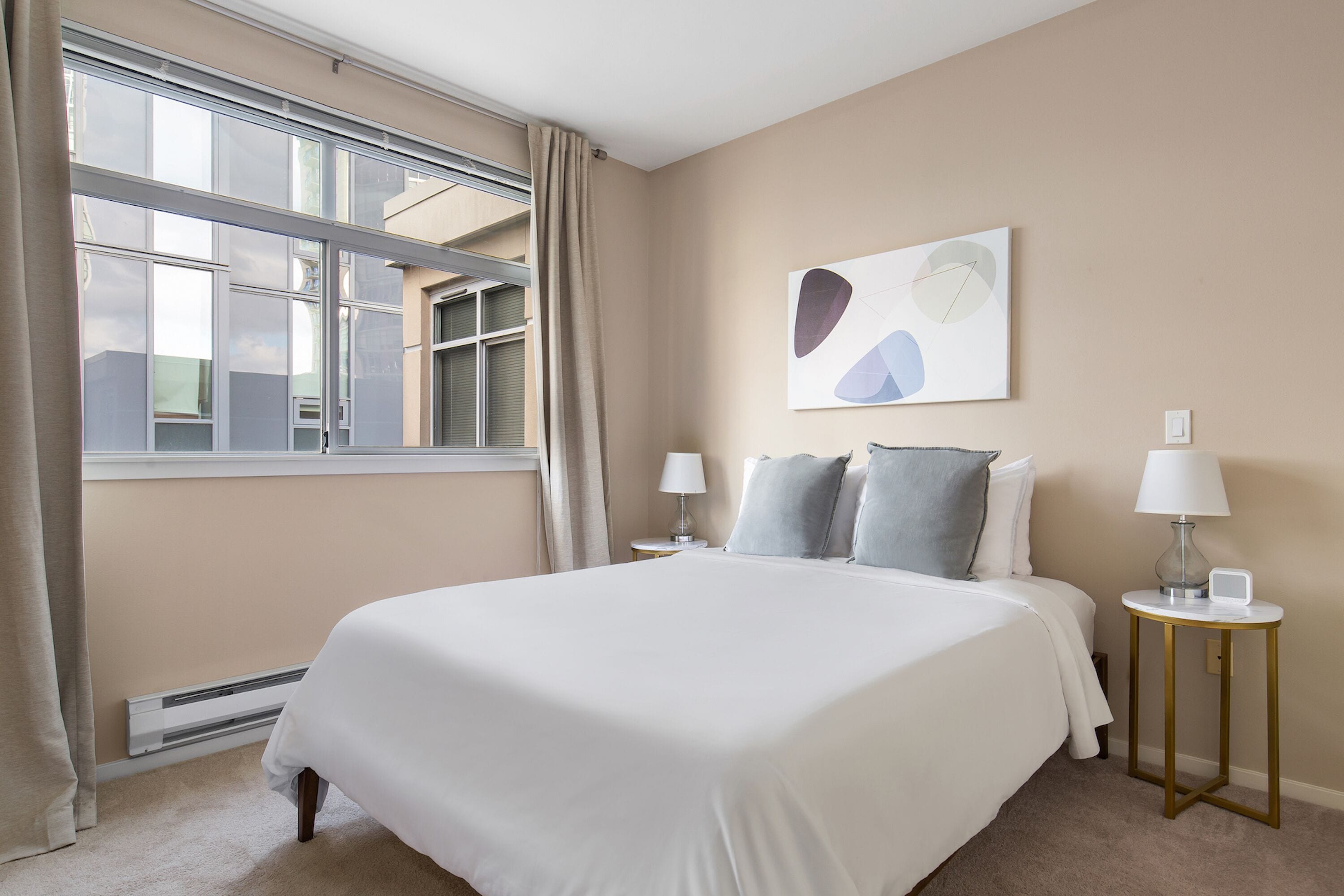 A neatly arranged bedroom with a large bed, two bedside tables with lamps, a window with curtains, and a wall-mounted abstract art piece.