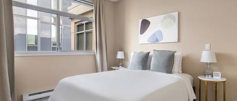 A neatly arranged bedroom with a large bed, two bedside tables with lamps, a window with curtains, and a wall-mounted abstract art piece.