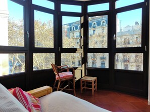 Una habitación con cama, una silla, un taburete pequeño y ventanas amplias que dan a edificios y árboles.