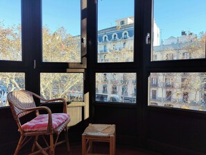 Una habitación con ventanas amplias, sillas de mimbre, un taburete pequeño y vistas a edificios.