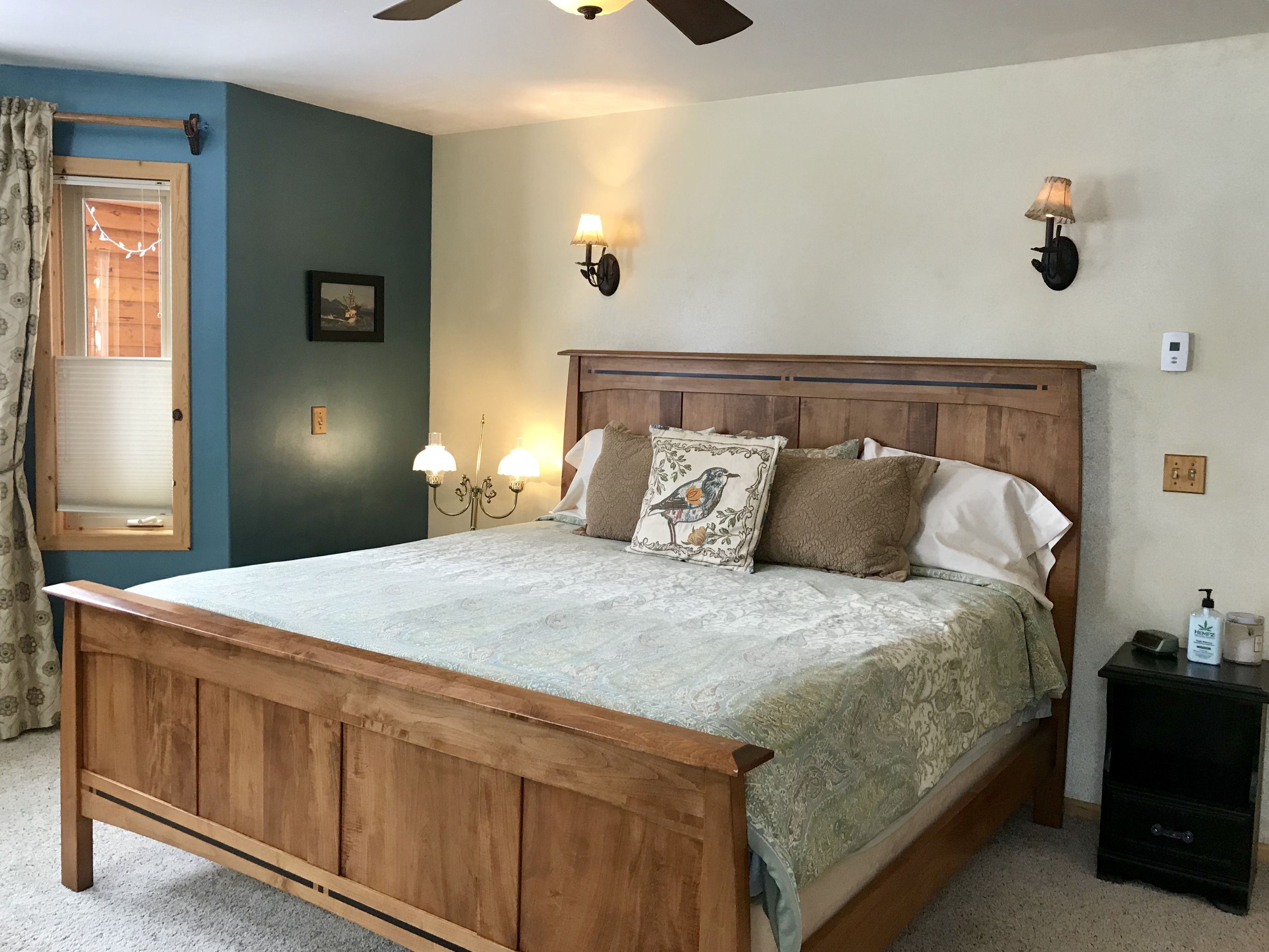 Un dormitorio con cama de madera, dos lámparas de pared, una mesita de noche y un ventanal con cortinas.