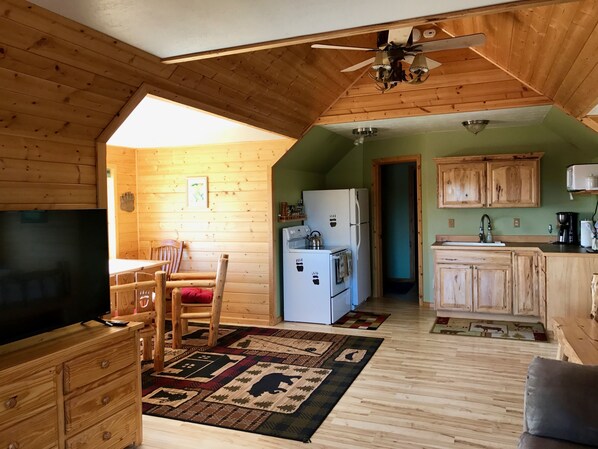 Una habitació acollidora d'estil cabana, amb sostre de fusta, ventilador de sostre, televisió, taula de dinar de fusta amb caires, zona de cuina amb armaris de fusta i una catifa amb dibuixos.