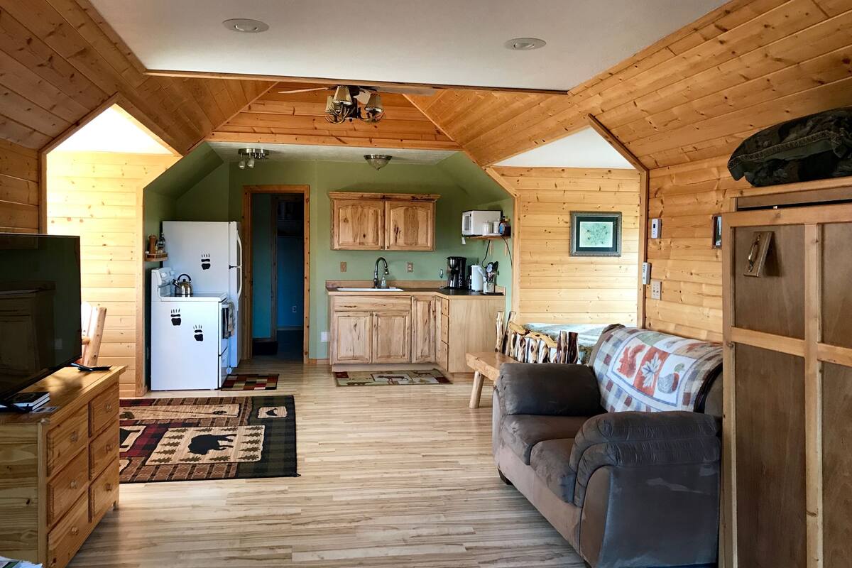 A cozy cabin interior with a wooden floor, a sofa, a TV, a kitchen with wooden cabinets, and a ceiling fan.