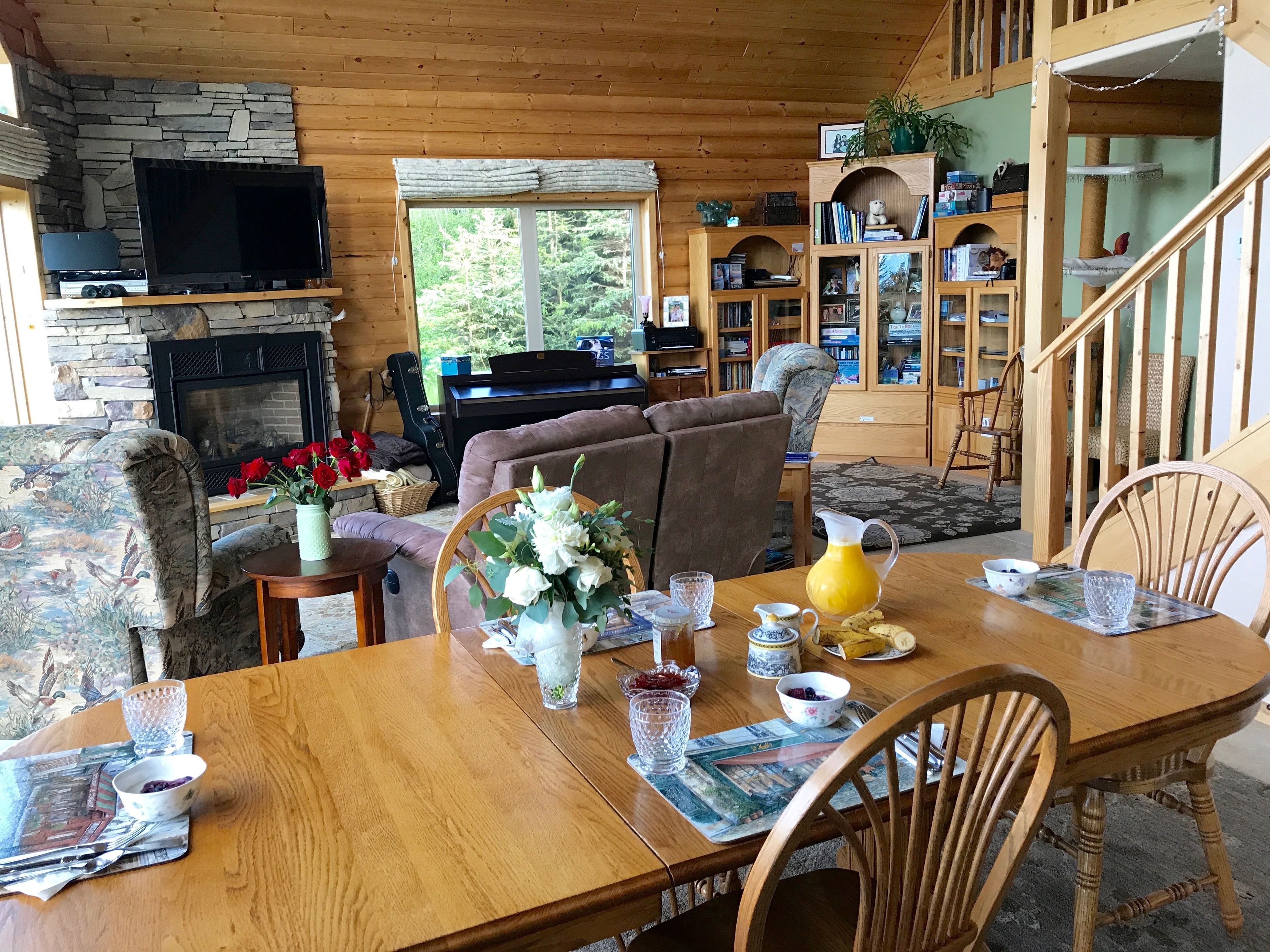 Interior acogedor de una cabaña con chimenea de piedra, un televisor, un piano y un comedor con mesa para dos.
