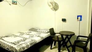 Une petite pièce simple, comprenant un lit, deux chaises, une table, un ventilateur et une prise de courant.