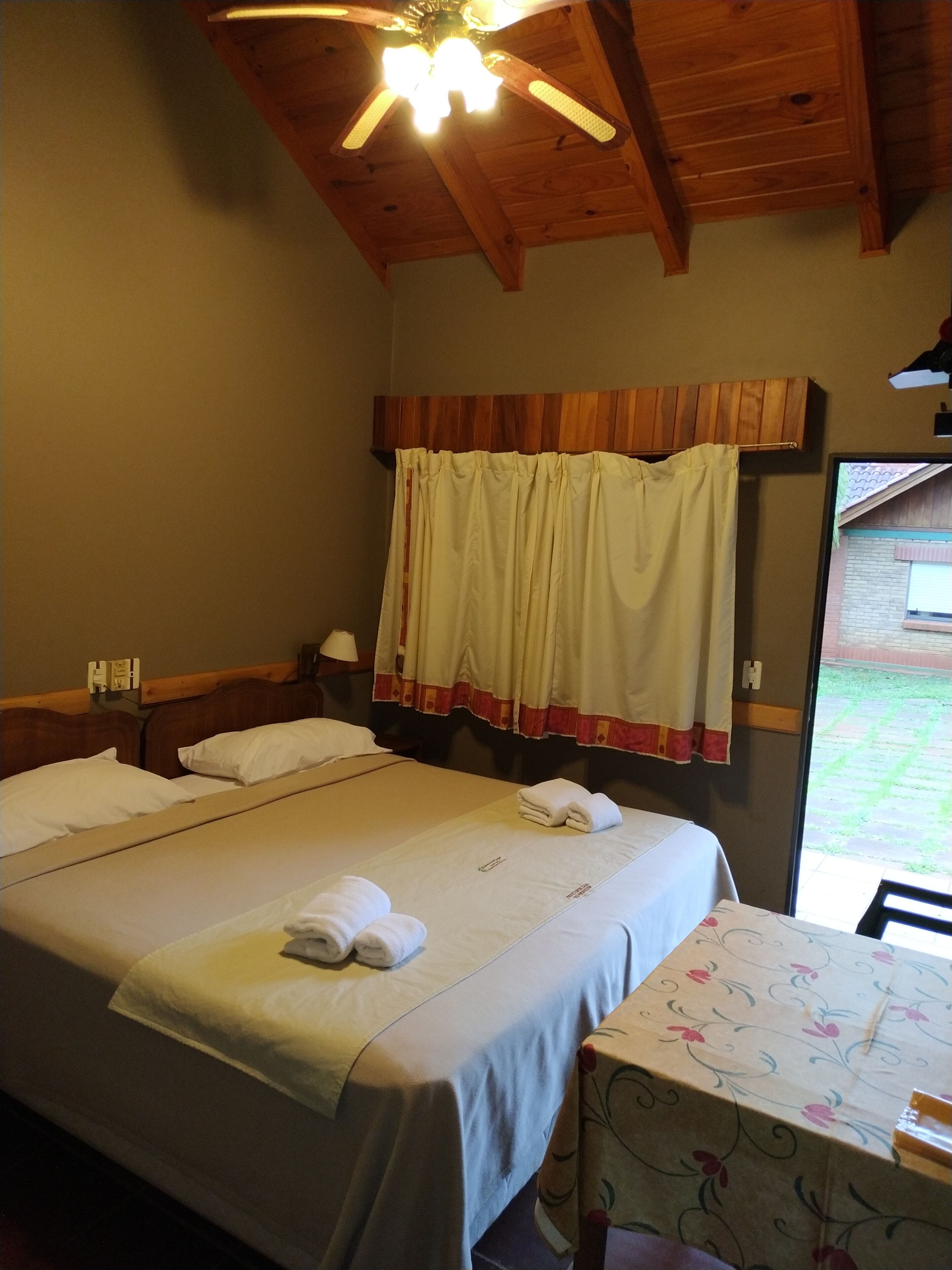 A hotel room with a bed, a table with a floral tablecloth, a ceiling fan, and a window with a view of the outside.