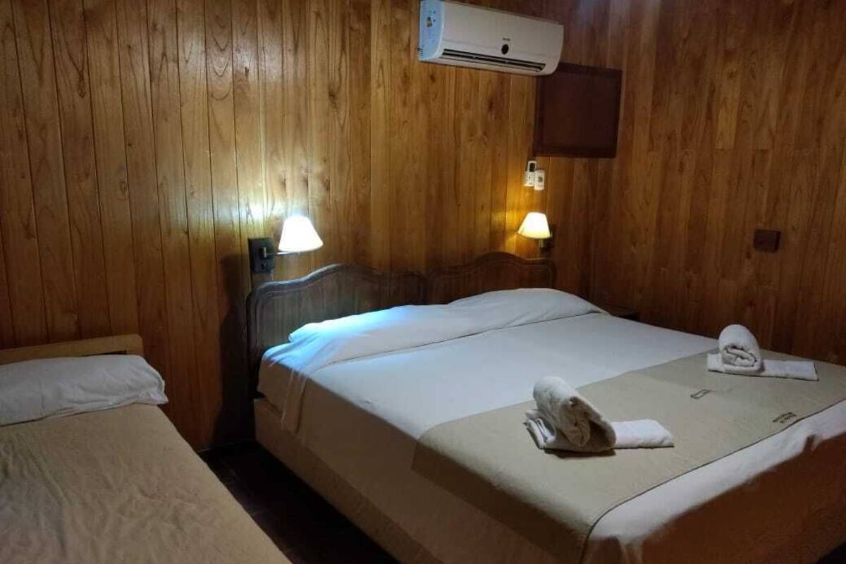 A room with wooden walls, a bed with a white bedspread, two bedside lamps, and an air conditioning unit.