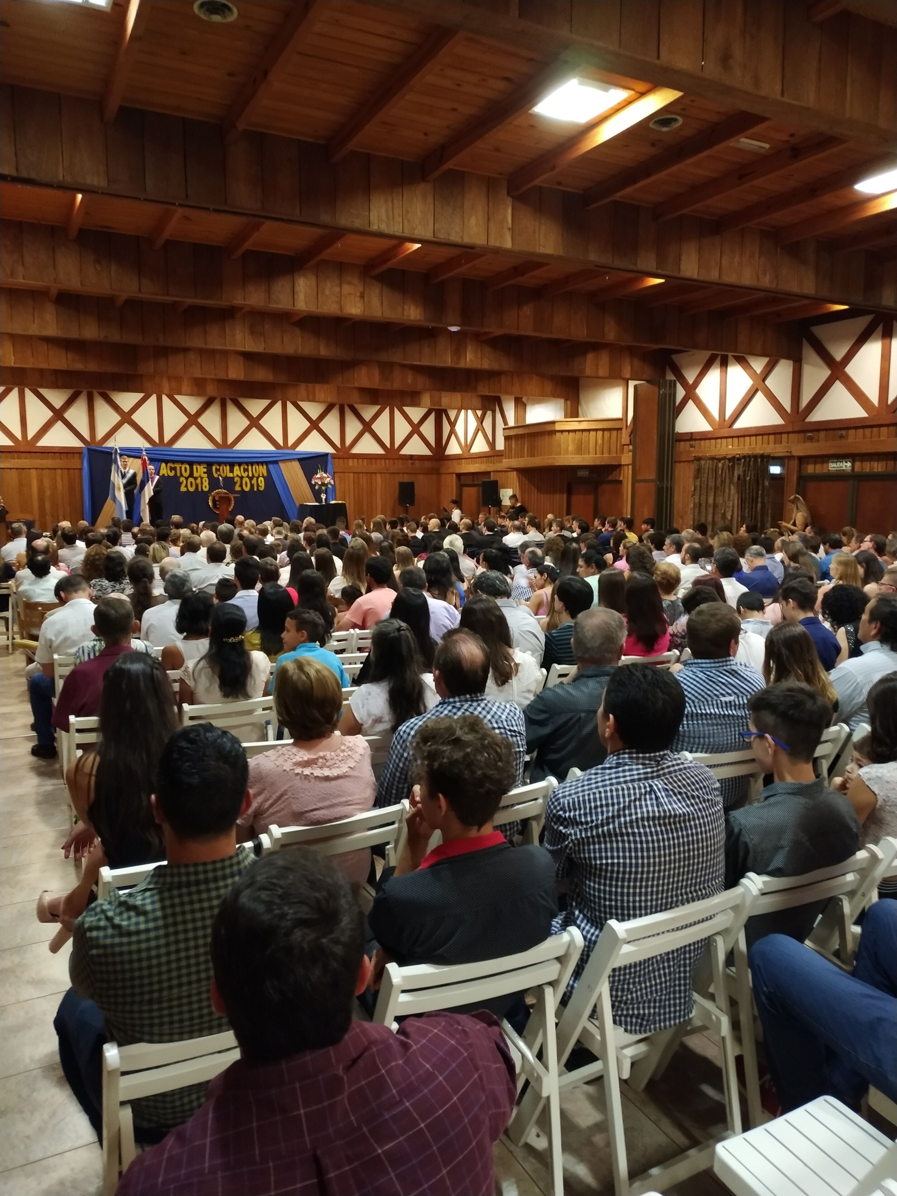 A large hall with wooden beams and a stage at the front. Attendees are seated in rows of chairs.