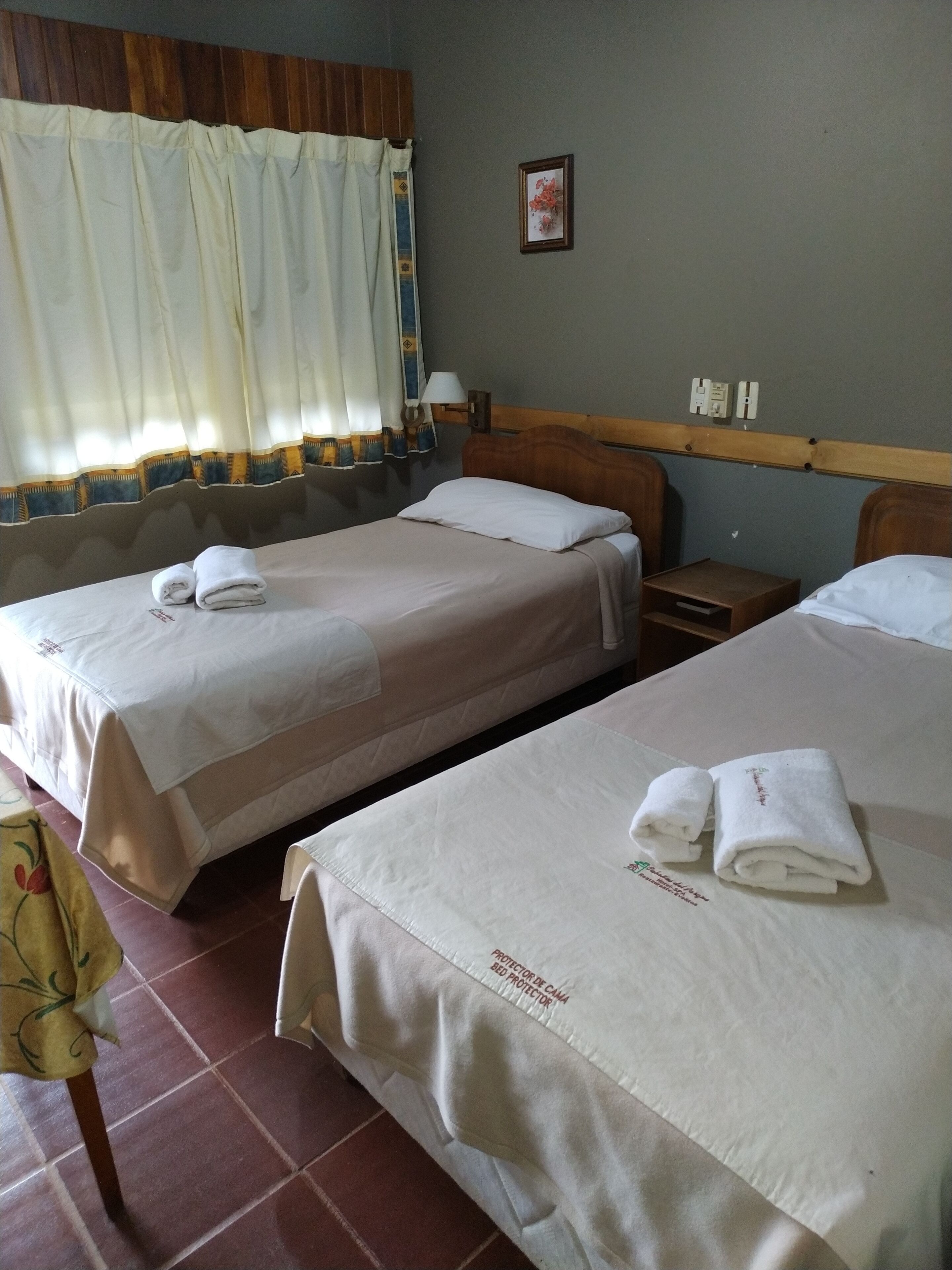 A hotel room with two beds, each with a white bedspread and a folded towel. There is a nightstand with a lamp, a wooden headboard, and a framed picture on the wall.