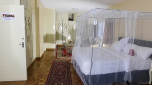 Una habitación de hotel con una cama cubierta por una mosquitera, piso de madera, un tapete rojo estampado y una puerta blanca.