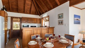 A dining area with a wooden table set for six, surrounded by chairs. A kitchen with wooden cabinets and a wooden ceiling is visible in the background. - Te Maiki Escape - Russell Holiday Home (Russell)