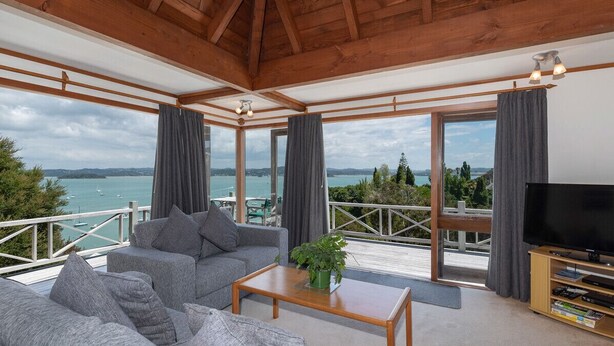 A modern living room with a large sofa, a coffee table, and a TV. The room has large windows offering a view of the sea and boats. - Te Maiki Escape - Russell Holiday Home (Russell)